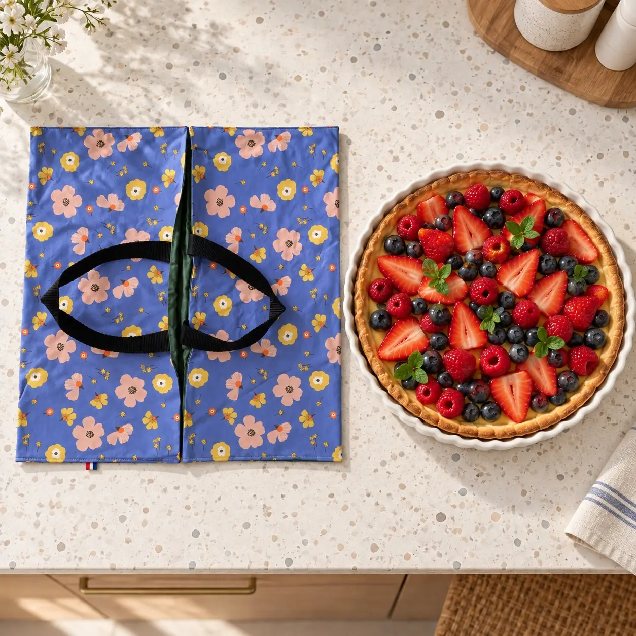Sac à tarte fabriqué en France Azalée – Fleuri bleu, imperméable et réversible 3 Sac à tarte bleu fleuri Timouny posé à côté d’une grande tarte aux fruits sur un plan de travail en terrazzo